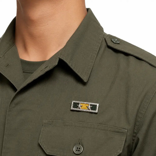 Homem em camisa verde-oliva com distintivo prateado, detalhes verde e amarelo, duas estrelas
