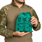 Homem com camisa bege segura saco verde KIT SOBREVIVÊNCIA NA SELVA, emblemas pretos.