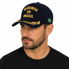 Homem de camiseta preta usando boné escuro Marinha do Brasil dourado com bandeira