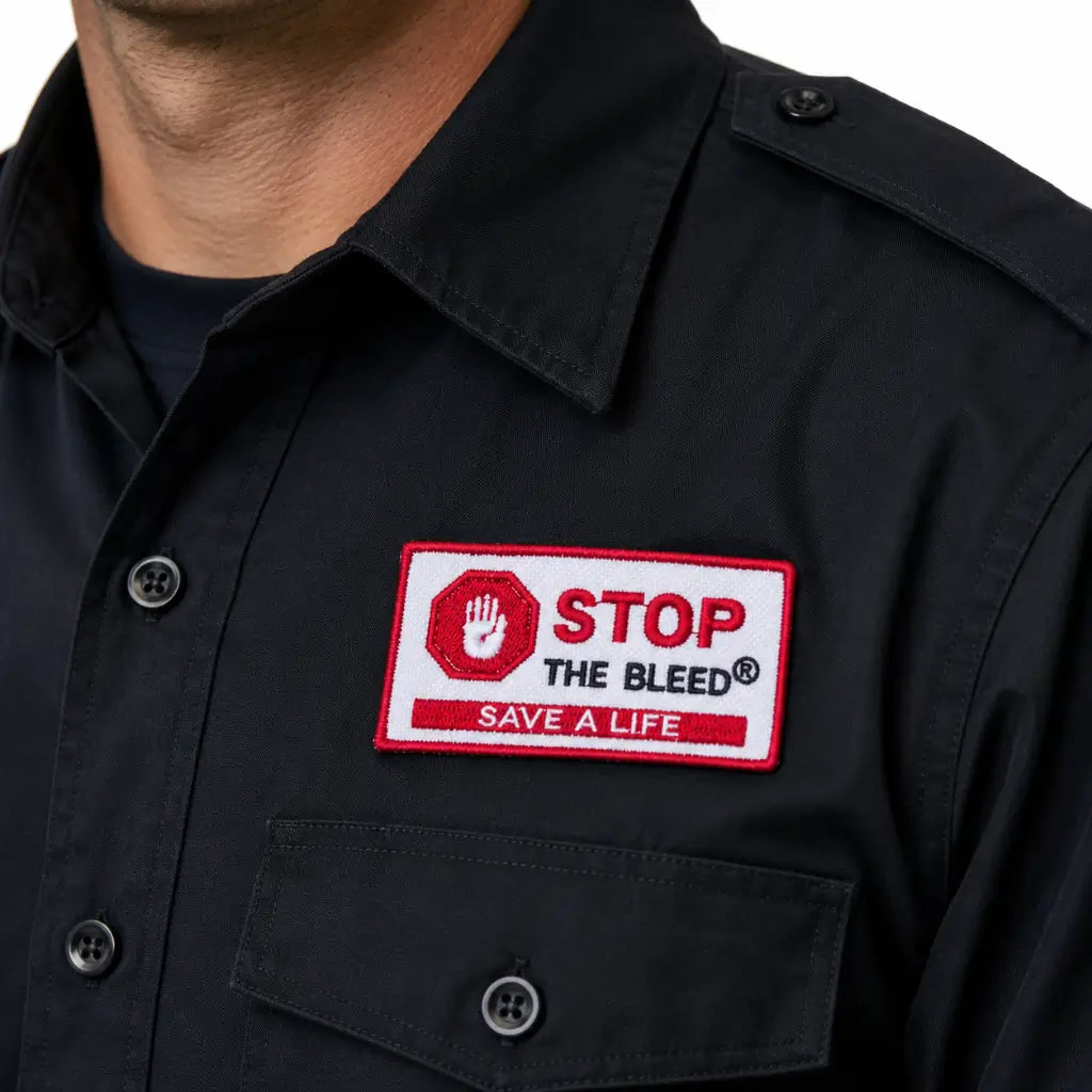Camisa preta com patch retangular branco; borda vermelha, mão e STOP THE BLEED