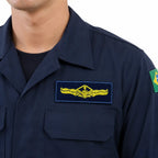 Pessoa em uniforme azul-marinho, emblema amarelo com asas e âncora no peito esquerdo; bandeira do Brasil na manga direita.